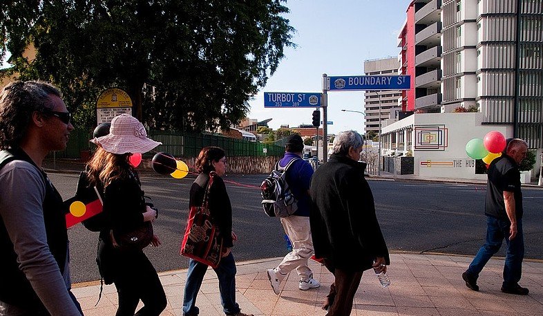 Walkers, one carrying the Aboriginal flag and others holding balloons, cross at the corner of Turbot Street and Boundary Street in Spring Hill.